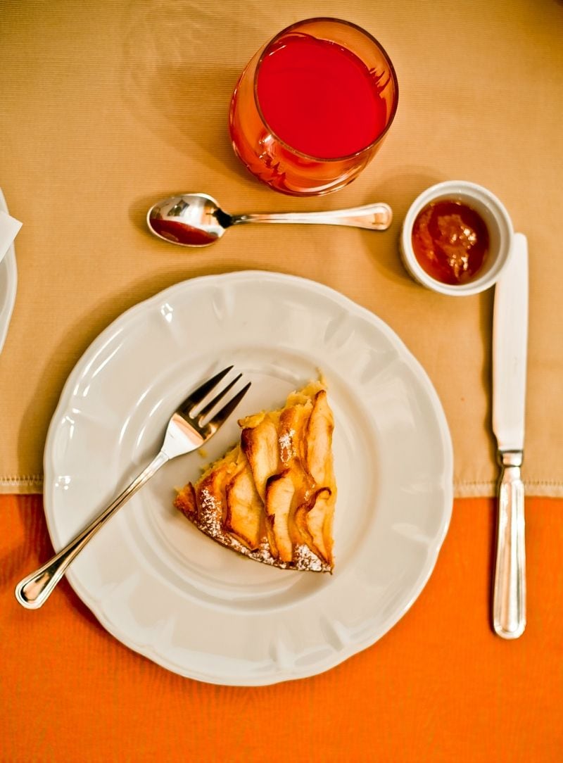 Una porción de pastel de manzana sobre un plato blanco, con un tenedor, una cuchara y un pequeño recipiente con salsa en una mesa.