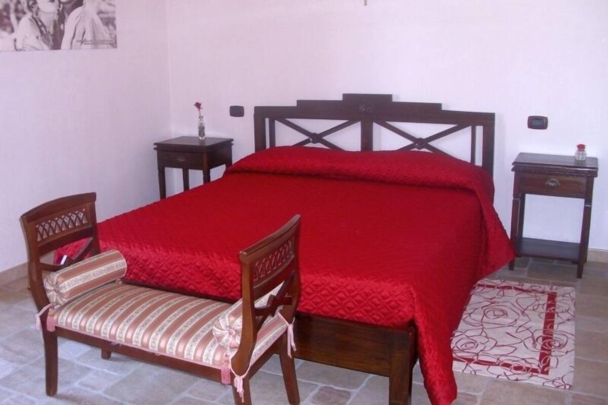 Une chambre à coucher avec un lit, une chaise en bois, deux tables de chevet, une croix accrochée au mur et un tableau encadré.