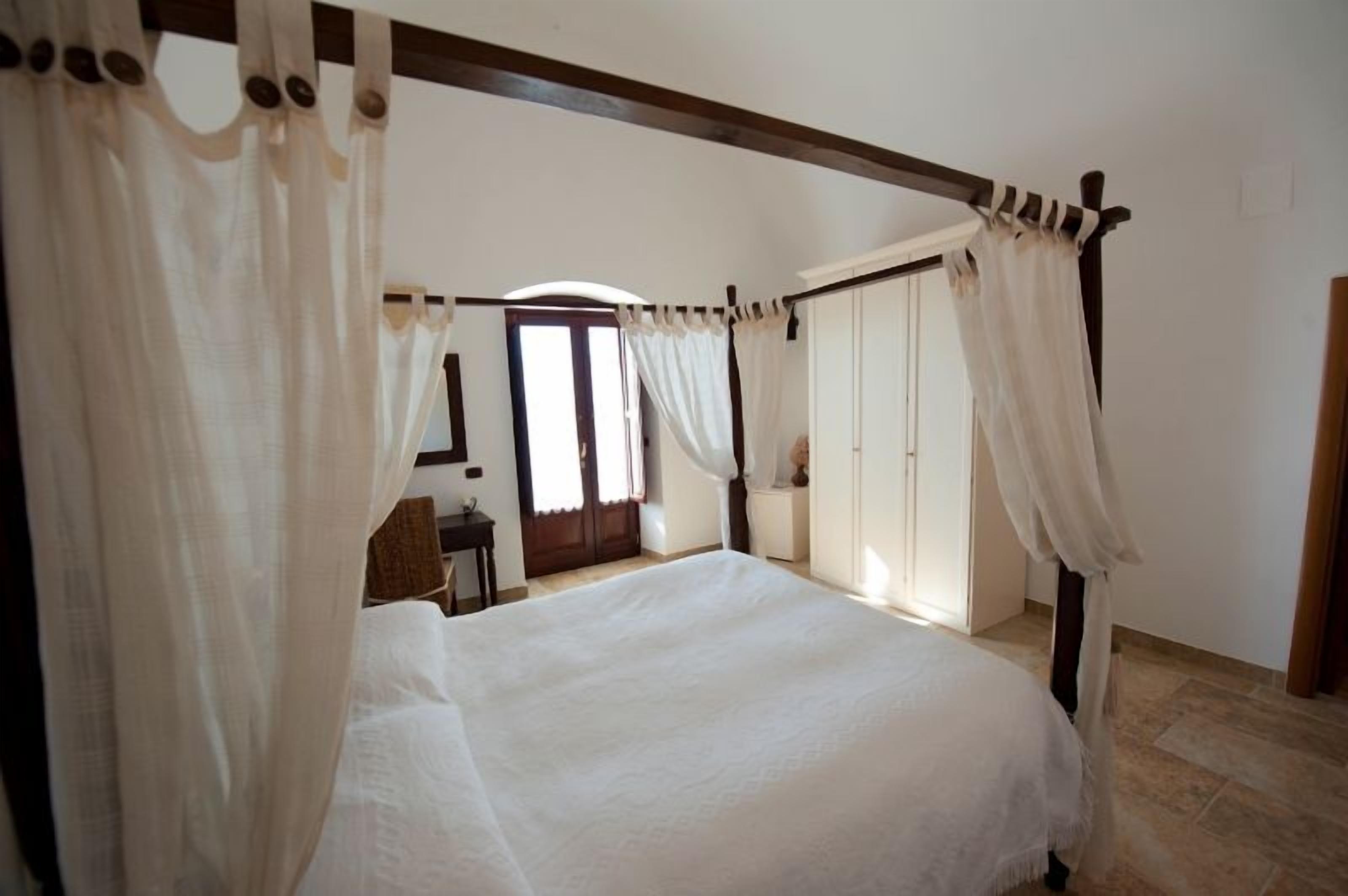 A four-poster bed with white linens, a wooden ceiling, and a view of a door and a small table in the background.