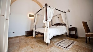 A four-poster canopy bed with a cross on the wall, a chair, and a small table beside it.