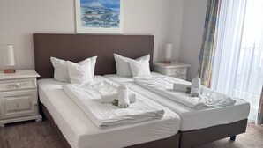 Un lit double avec du linge de lit blanc, une tête de lit marron et un tableau représentant un paysage accroché au mur.