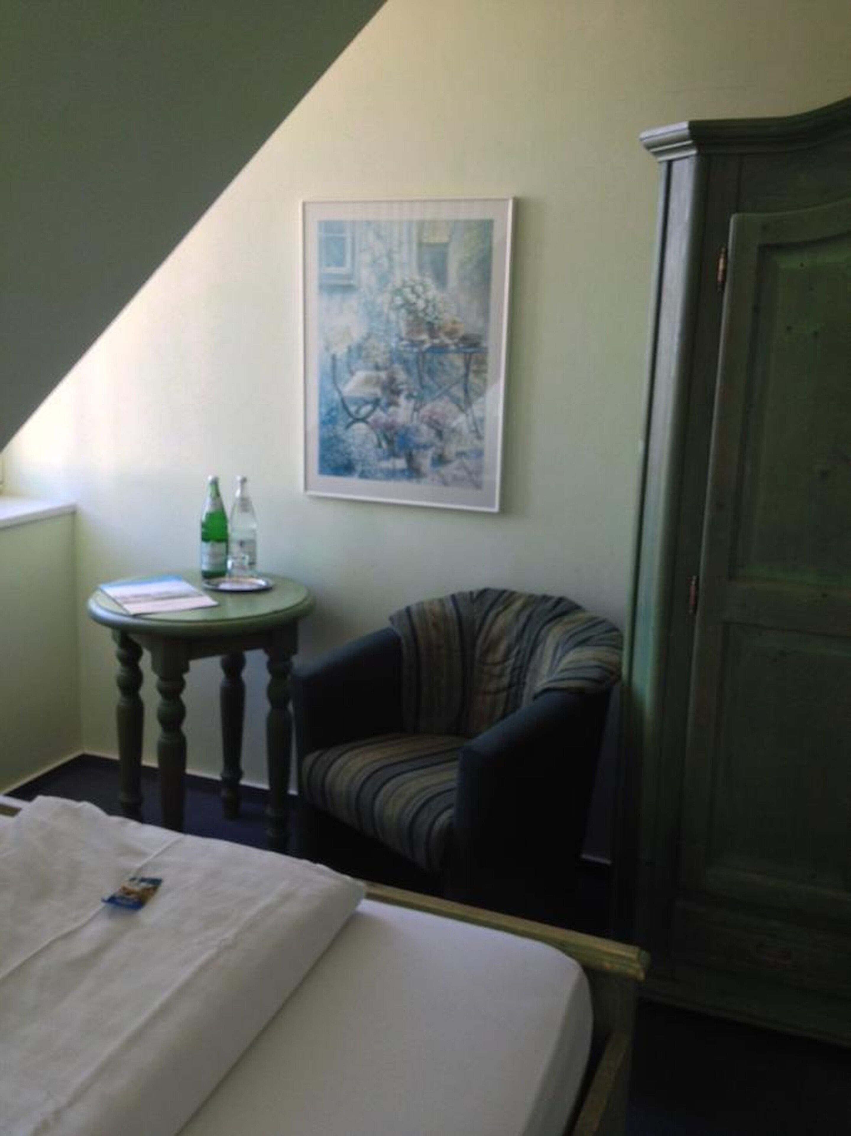 A room with a bed, a chair, a small table with a bottle, and a painting on the wall.