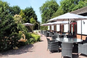 An outdoor patio area with round tables and chairs, surrounded by lush greenery and trees. - The Stanwick Hotel (Wellingborough)
