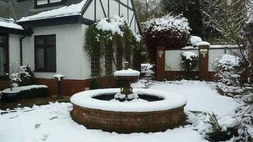 A snow-covered garden with a circular fountain, a brick wall, and a house with a white facade and dark trim.