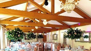 Un salón de banquetes con vigas de madera, candelabros y centros de mesa florales. Las mesas están dispuestas con manteles blancos y cubresillas rosas.
