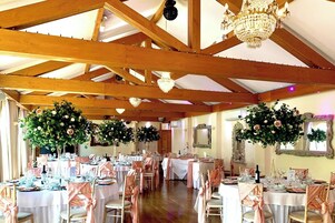 Un salón de banquetes con vigas de madera, candelabros y centros de mesa florales. Las mesas están dispuestas con manteles blancos y cubresillas rosas.