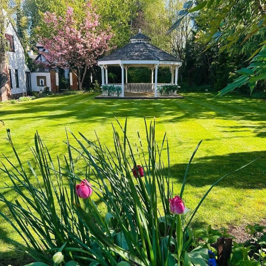 Un jardín cuidado con césped, árboles y plantas en flor, donde se encuentra un gazebo.