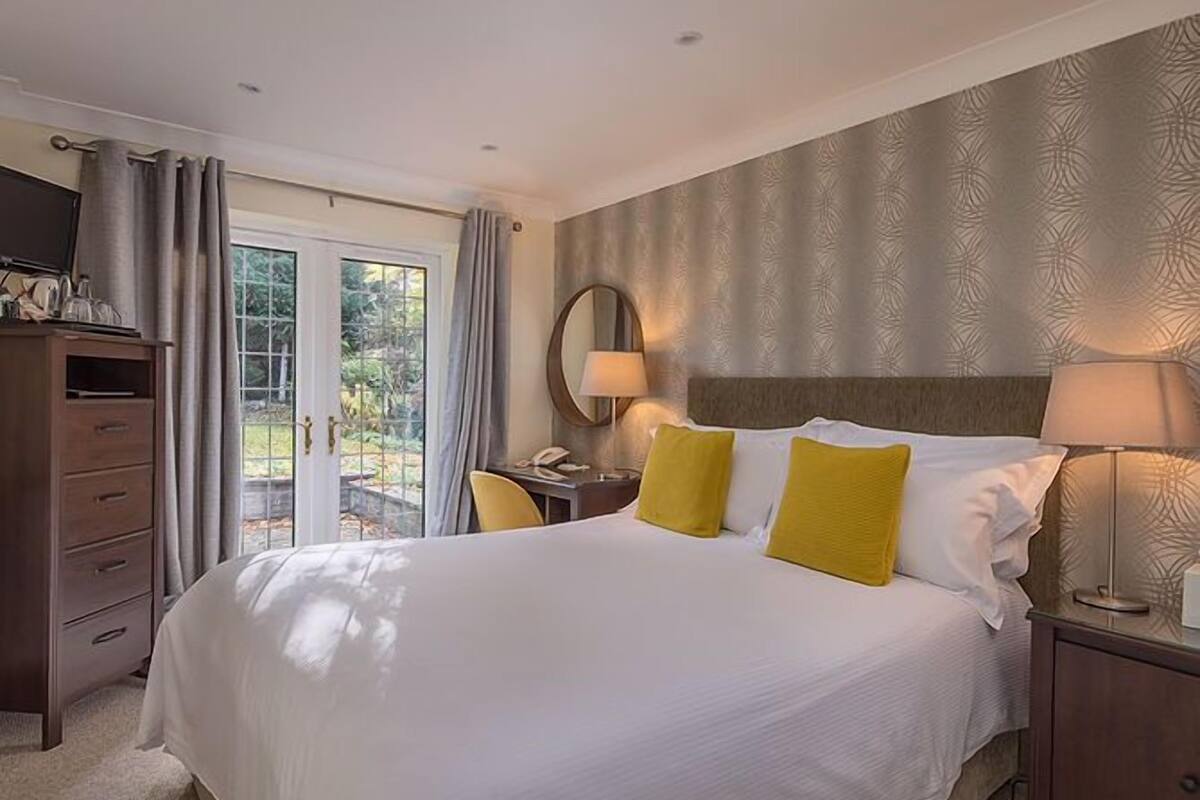 A hotel room with a large bed, a desk with a lamp, a chair, and a wardrobe.