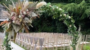 Un salón de bodas al aire libre con filas de sillas blancas y un arco floral.