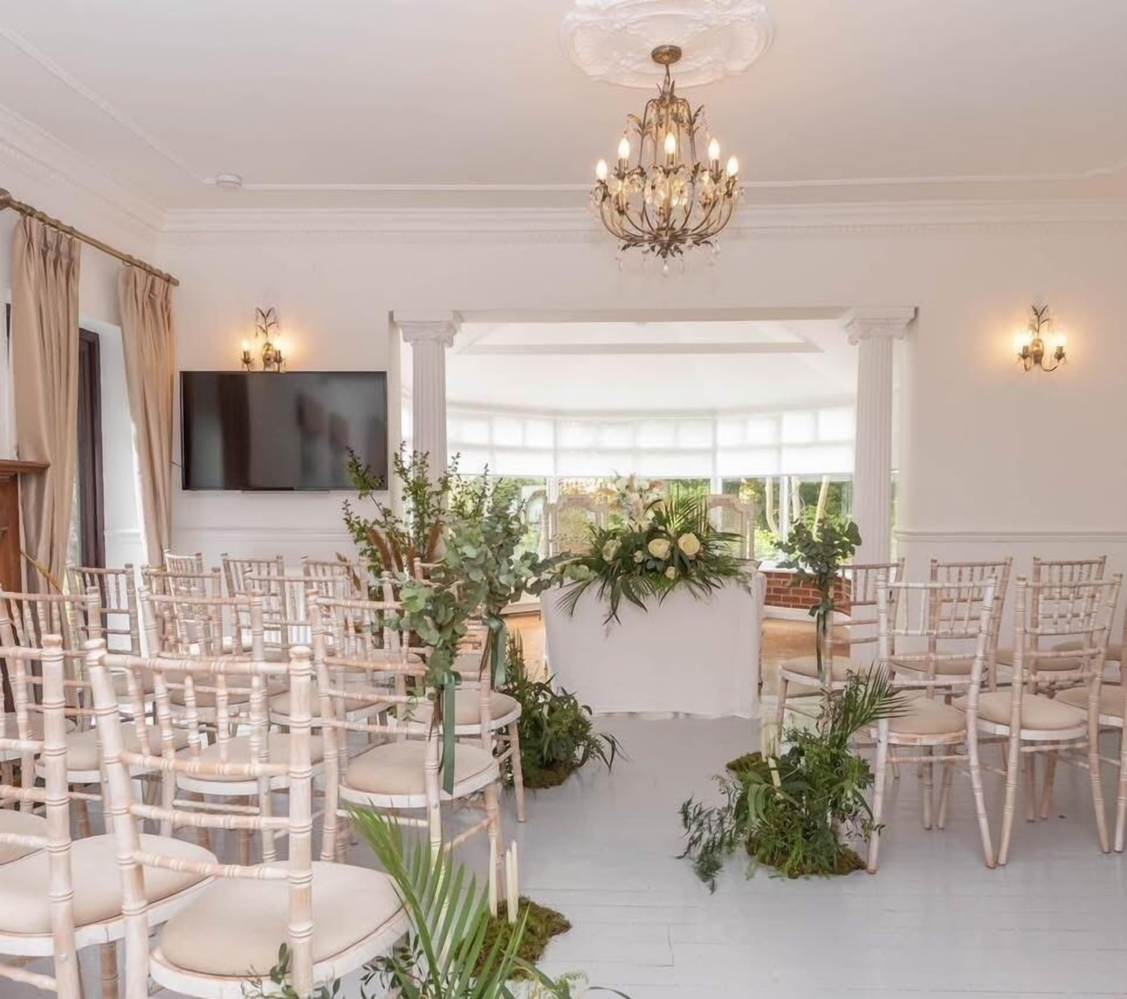 A formal indoor setting with rows of white chairs, a central floral arrangement, and a chandelier.
