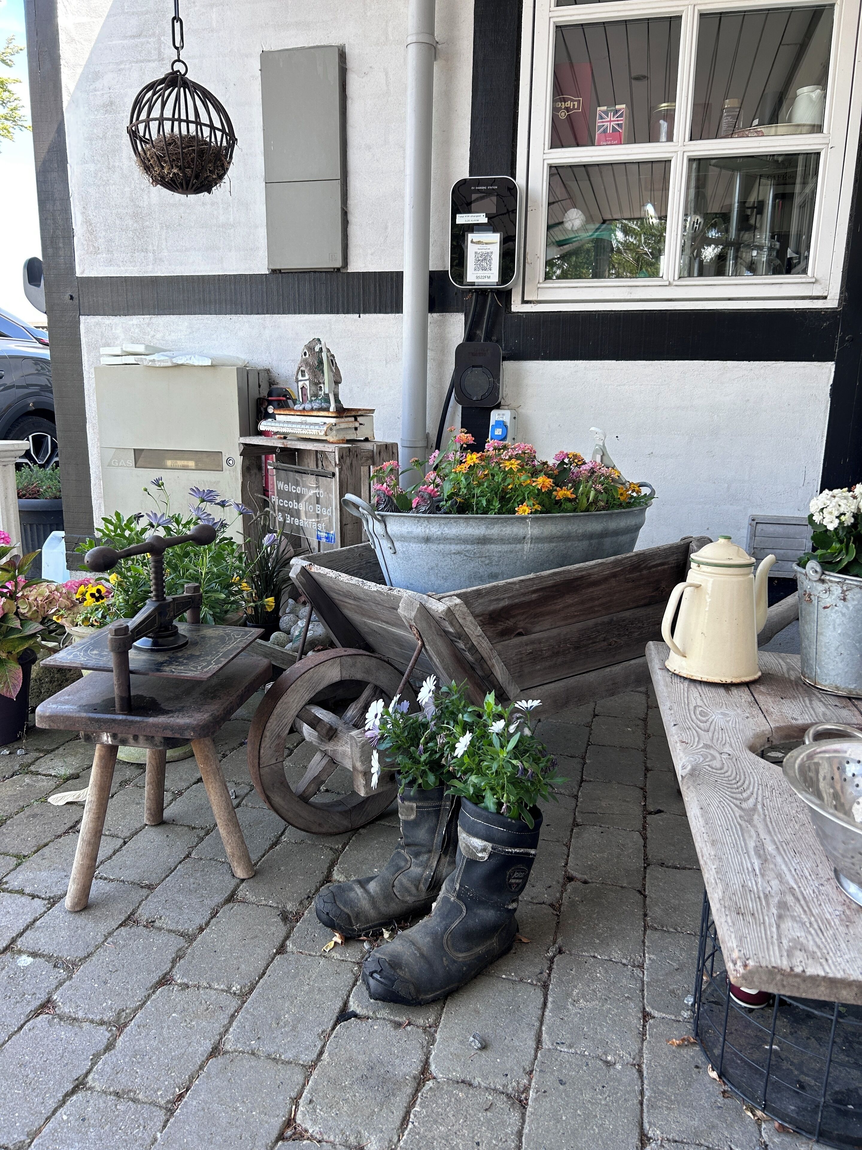 A rustic outdoor seating area with a wooden table, a metal bucket with flowers, a vintage coffee pot, and a pair of boots.