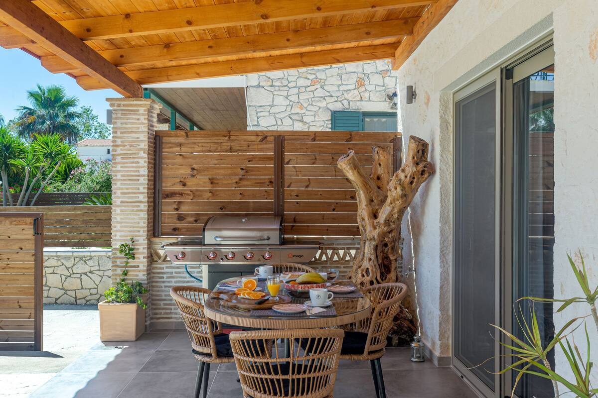 Eine Terrasse im Freien mit einer Steinwand, einer Holzbalkendecke und einem Grill. Wicker-Stühle und ein Tisch sind gedeckt mit Tellern, Tassen und einer Banane.