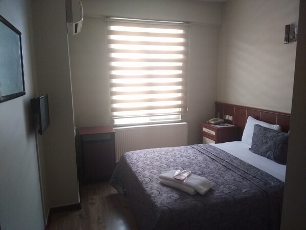 A hotel room with a bed, a bedside table with a telephone, a window with blinds, and a radiator. - Yeni Hotel (Malatya)