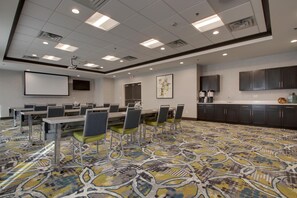 A conference room with a large table, chairs, a projector screen, and a kitchen area. - Hampton Inn & Suites Knightdale Raleigh (Raleigh)