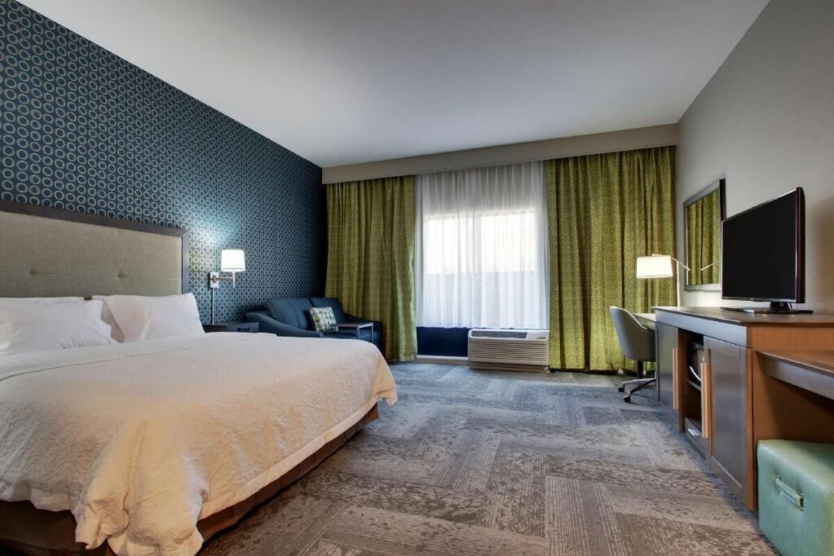 A hotel room with a large bed, a desk with a television, a chair, and a window with curtains.