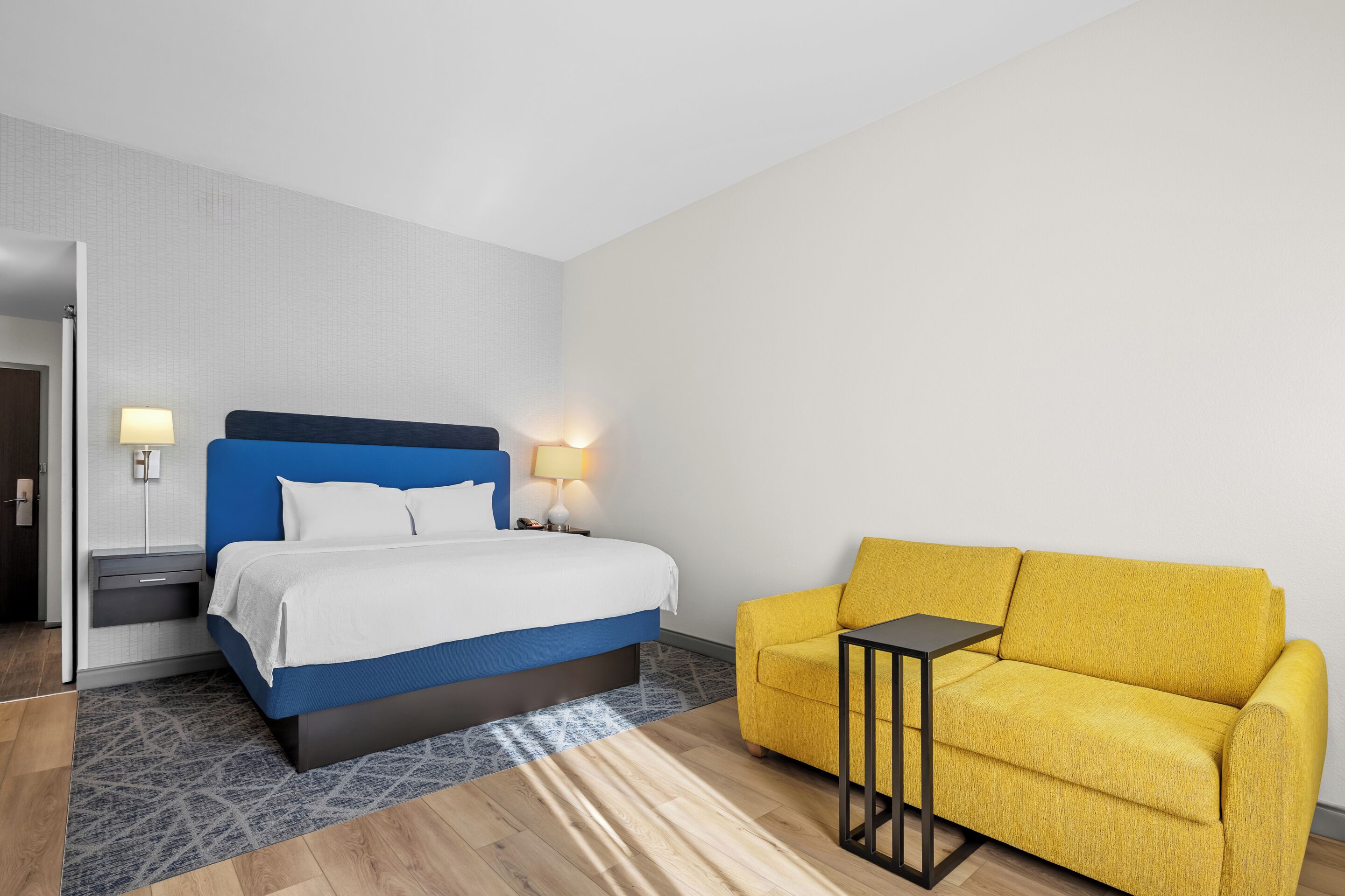 A hotel room with a bed, a sofa, a nightstand, and a lamp.