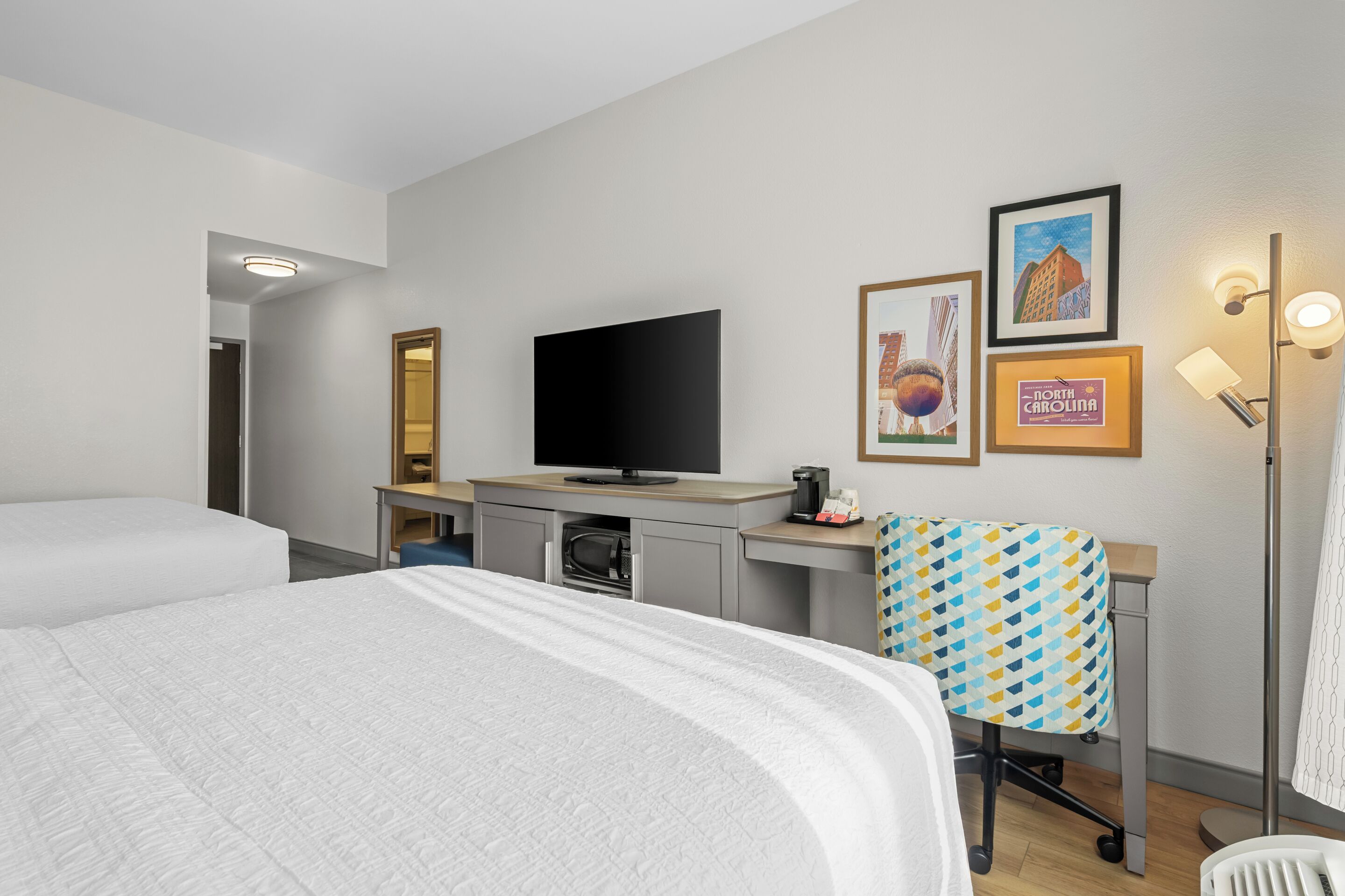 A hotel room with a bed, a desk with a television, a microwave, and a chair. There are framed pictures on the wall.