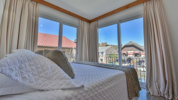 A hotel room with a large bed, beige curtains, and a view of buildings through large windows. - Hotel Micheline (Gramado)