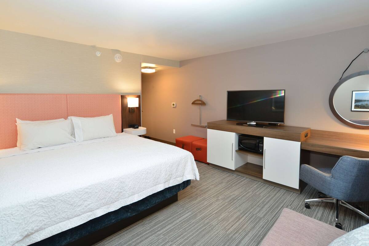 A hotel room with a bed, a desk with a TV, a chair, and a small orange ottoman.