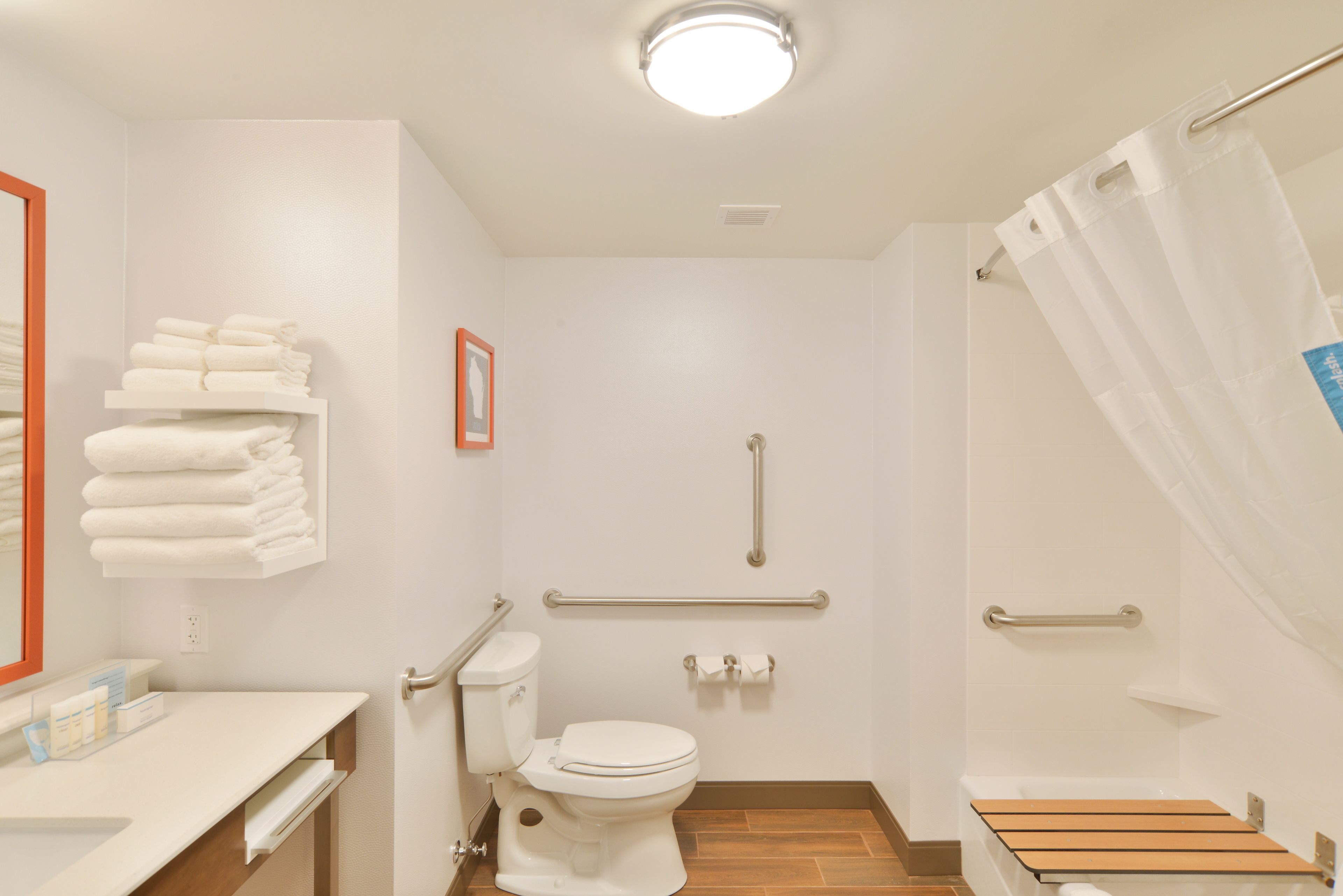 Un baño pequeño y limpio con inodoro, lavamanos y zona de ducha. Cuenta con barras de apoyo y un banco de madera.