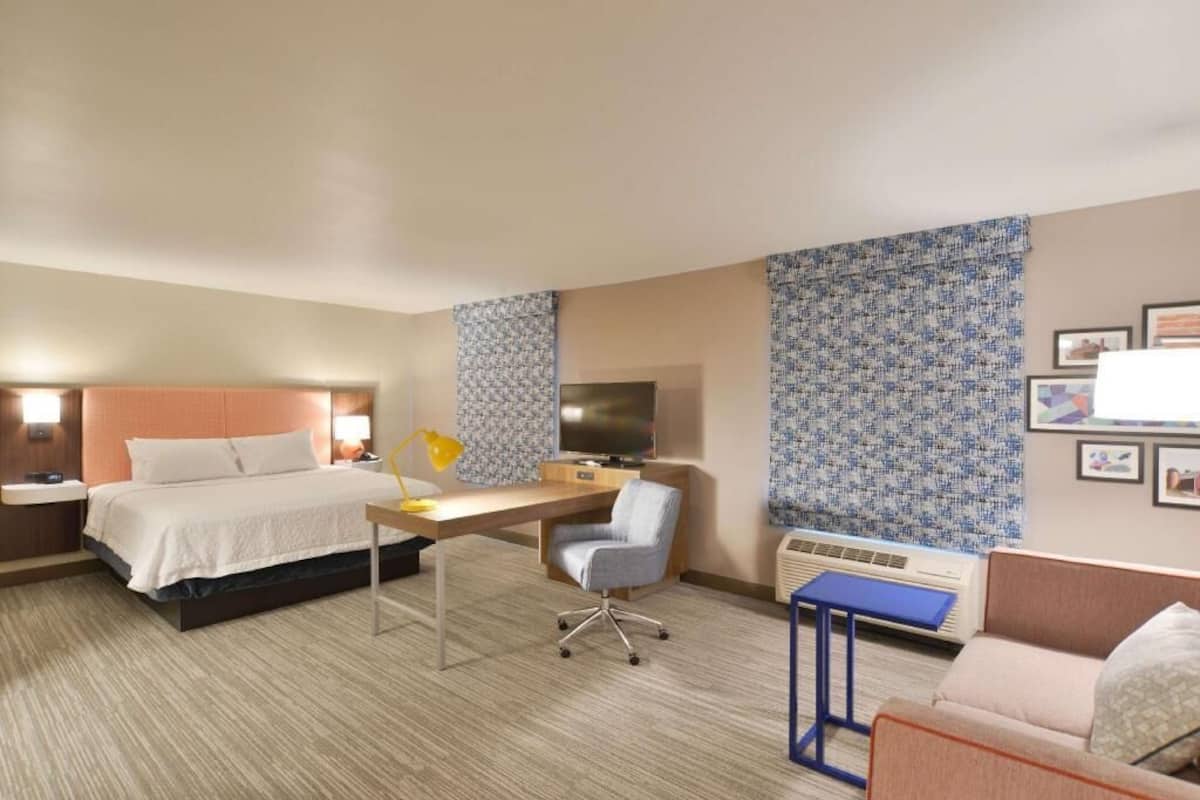 A hotel room with a bed, desk, chair, TV, air conditioning unit, and framed pictures on the wall.