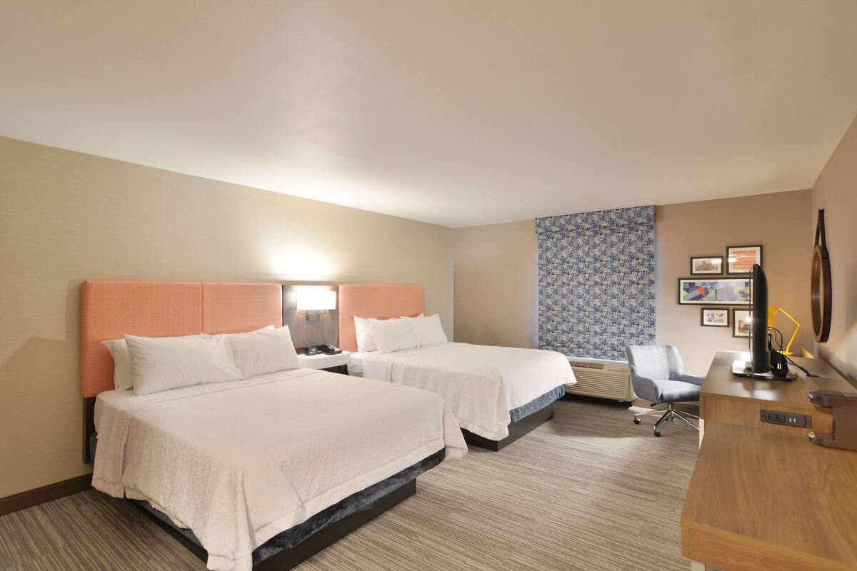 A hotel room with two beds, a desk, a chair, and a patterned wall.