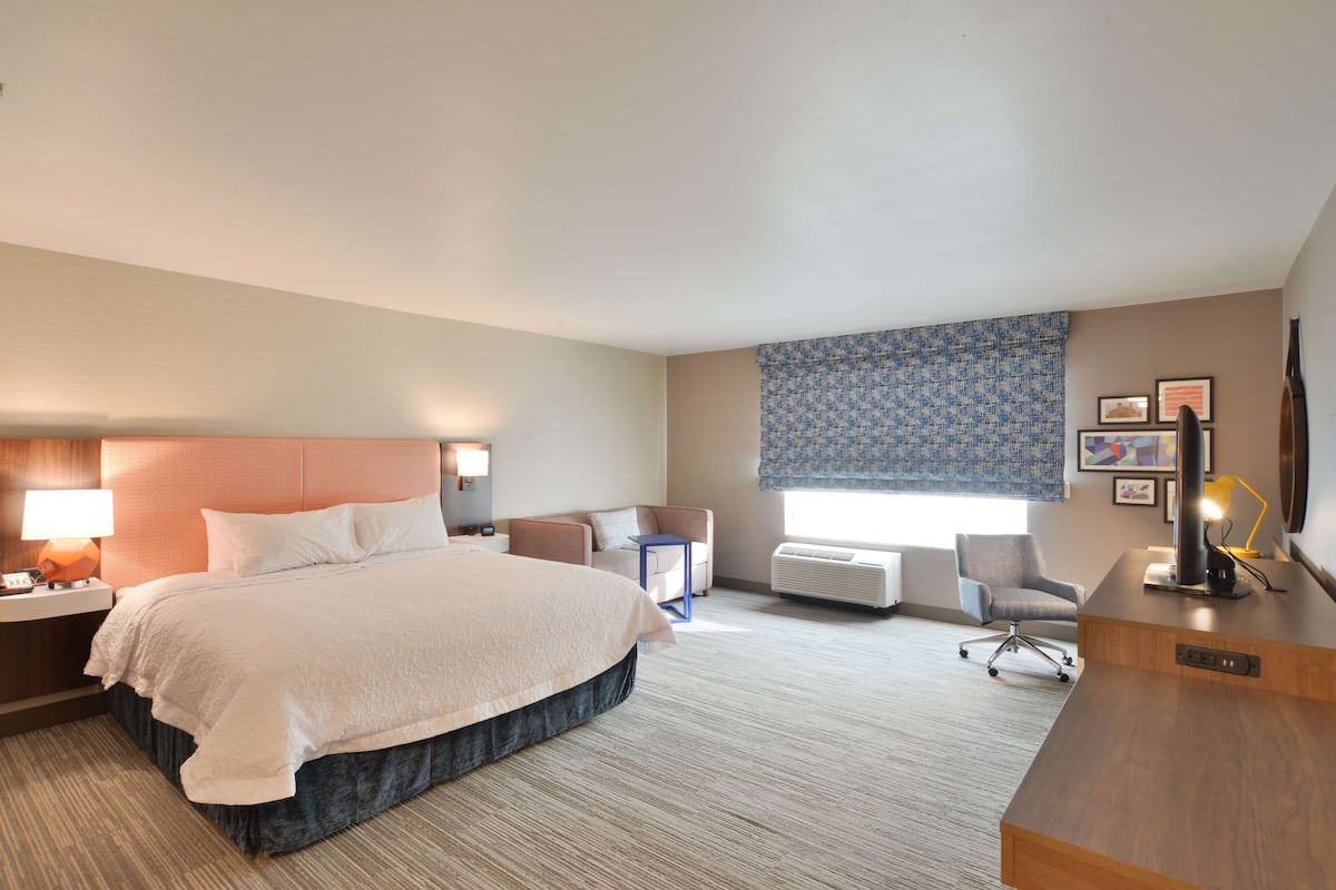 A hotel room with a large bed, a desk, a chair, a small table, and a window with patterned curtains.