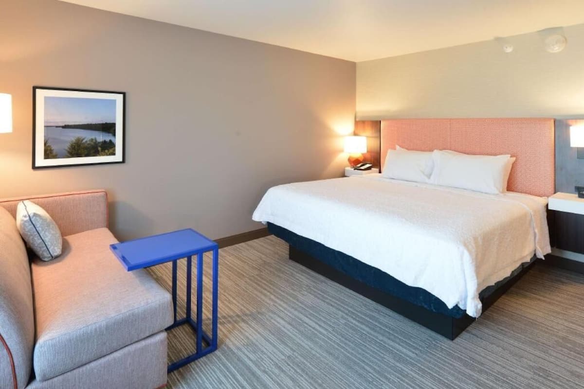 Une chambre d’hôtel avec un lit, une chaise, une petite table, une table de chevet, une lampe et un tableau encadré au mur.