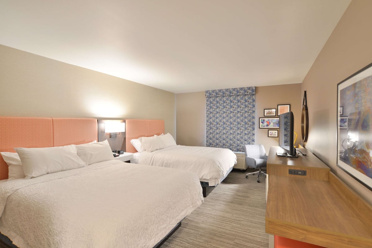 A hotel room with two beds, a desk, a chair, and a large framed picture on the wall.