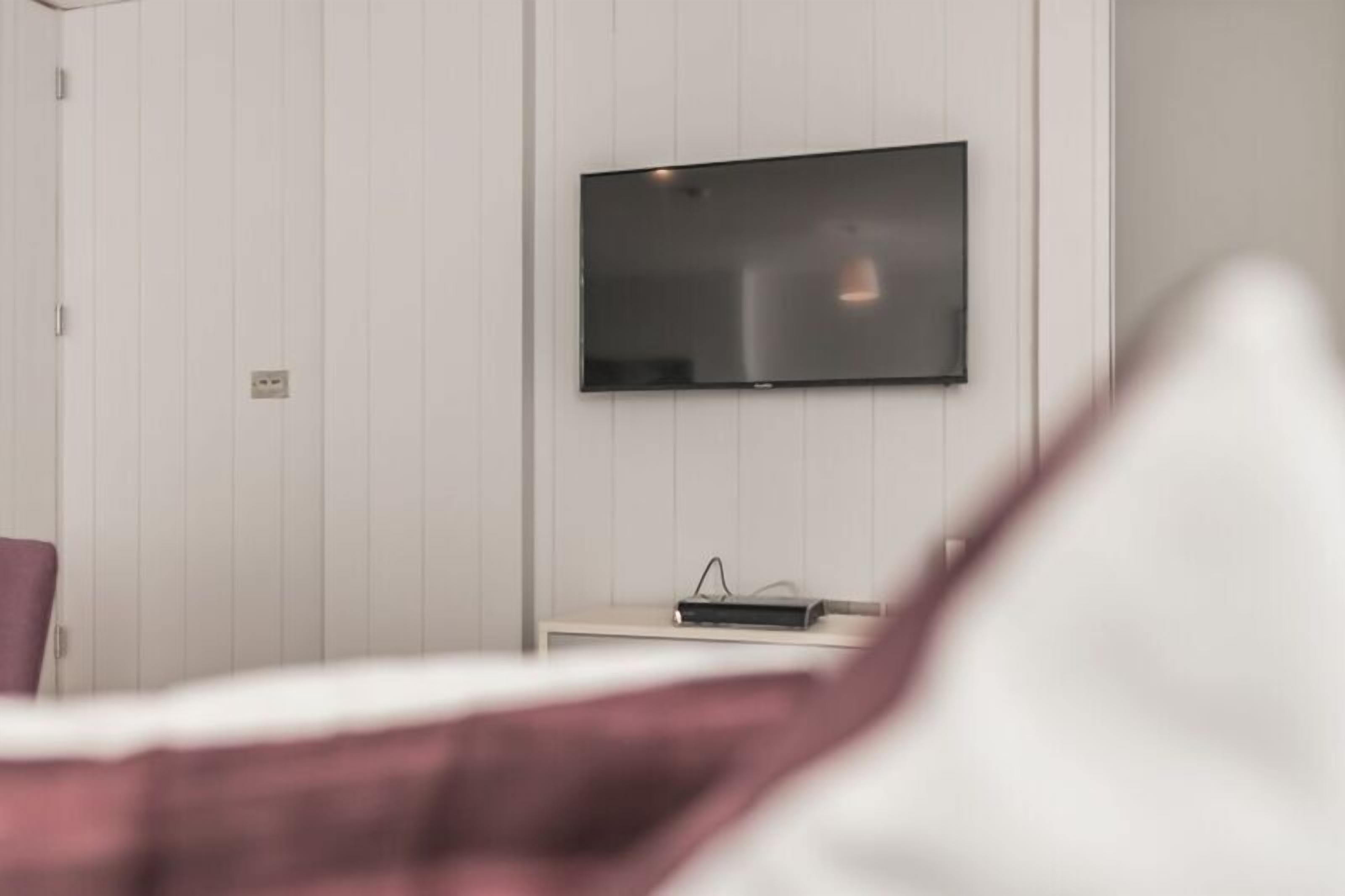 A modern hotel room with a flat-screen TV mounted on the wall, a bedside table with a remote control, and a bed with a white and purple bedspread.