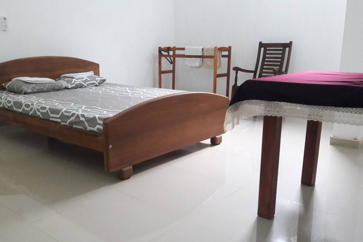 Ein Schlafzimmer mit einem hölzernen Bett, einem kleinen Tisch mit Spitzenbettdecke, einem Sessel und einem an der Wand befestigten Regal.