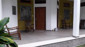 Een overdekte veranda met een houten deur, twee houten stoelen, een bloempot en een witte emmer.