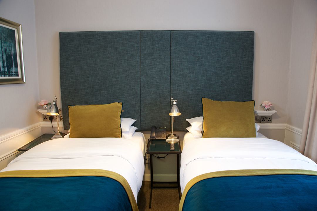A hotel room with two beds, a large grey headboard, bedside tables with lamps, and a small nightstand with a phone.