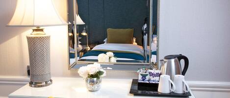 A hotel room with a bed, a bedside table with a lamp, a mirror, a vase with flowers, and a tray with coffee cups and a kettle.