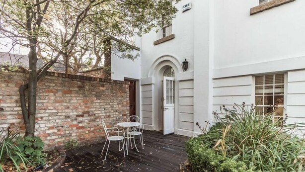 Un edificio blanco con un muro de ladrillo, un pequeño patio con una mesa y sillas, y una puerta con contraventanas.