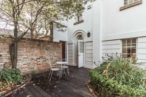 Een wit gebouw met een bakstenen muur, een klein terras met een tafel en stoelen, en een deur met luiken.