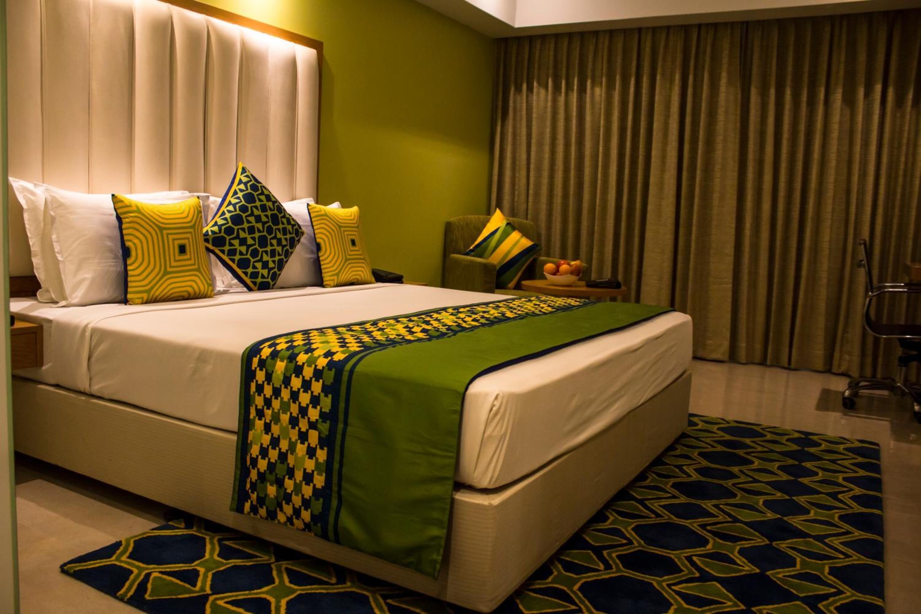 Habitación de hotel con una cama grande, colcha y alfombra estampada en verde y amarillo, una mesita de noche con lámpara, y vistas al exterior a través de cortinas.
