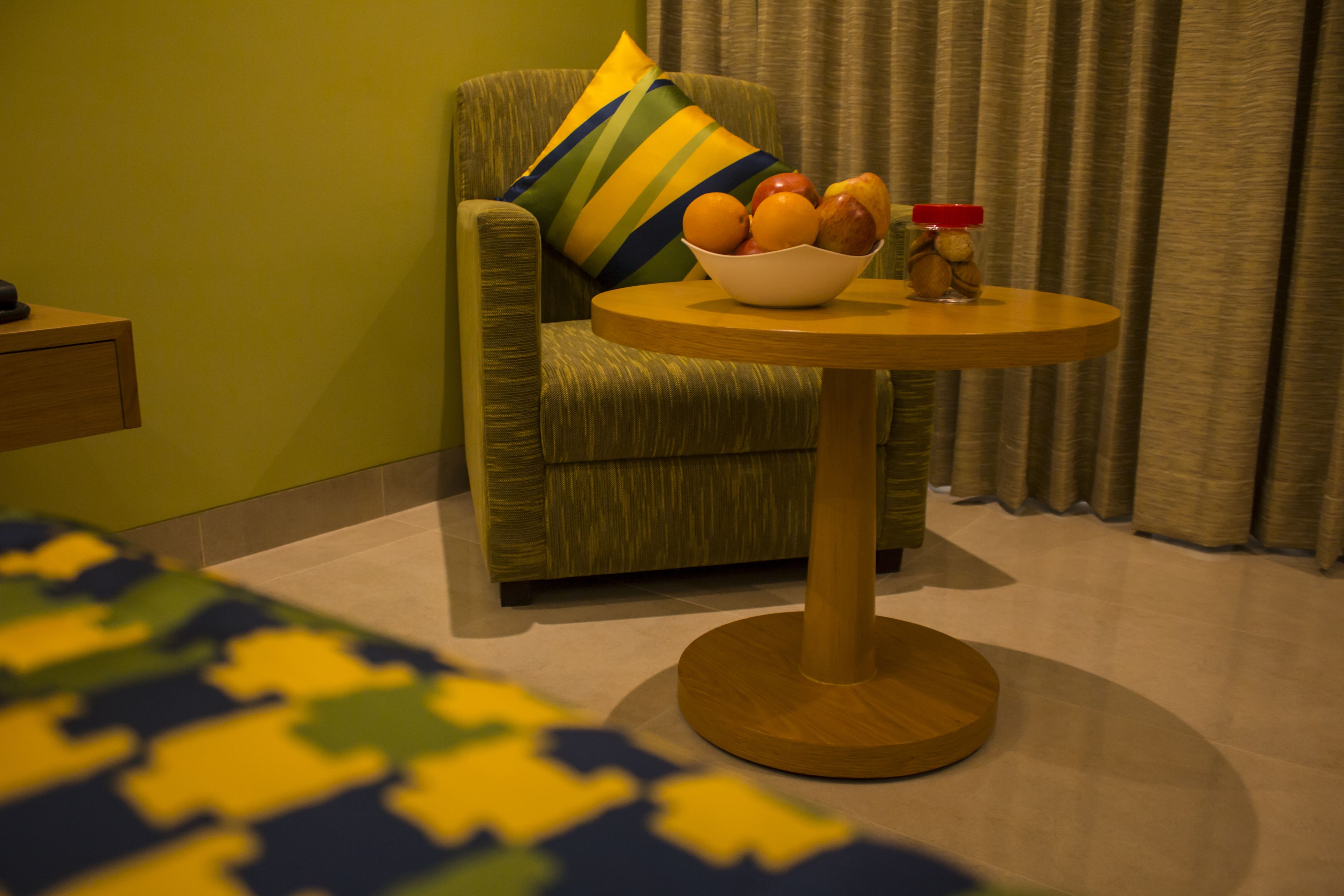 Una habitación de hotel con un sillón verde, una mesa redonda de madera con un frutero y una cama con colcha estampada.