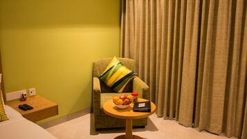 A room with a green armchair, a small round table with a bowl of fruit and a remote control, and a curtain.