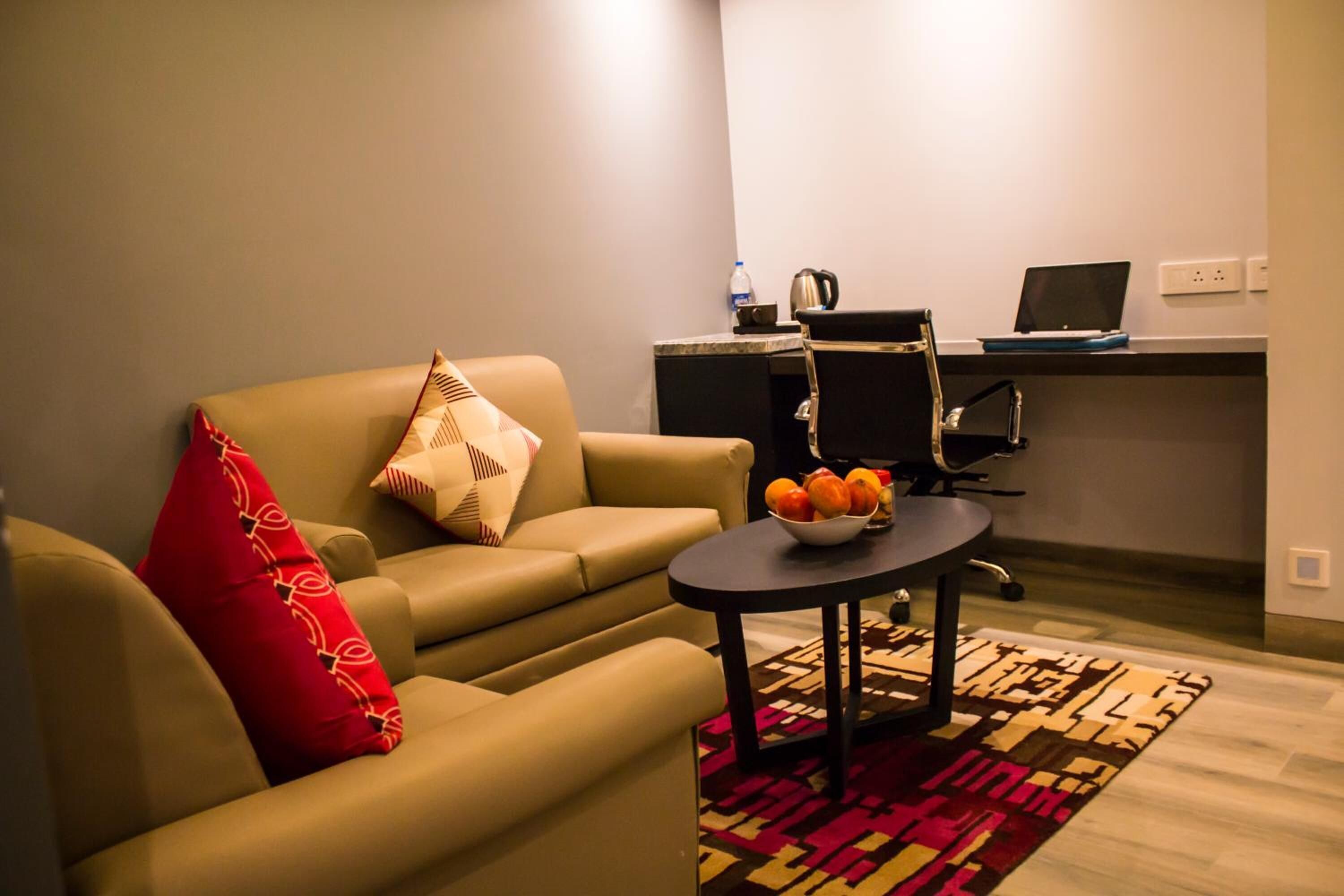 A living room with a sofa, a small round table with a fruit bowl, a red pillow, and a patterned rug. There is a kitchenette area with a laptop and a kettle.