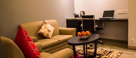 A living room with a sofa, a small round table with a fruit bowl, a red pillow, and a patterned rug. There is a kitchenette area with a laptop and a kettle.