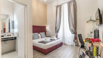 Habitación de hotel con cama, mesita de noche, espejo, zona de ducha y una pequeña mesa con un florero.