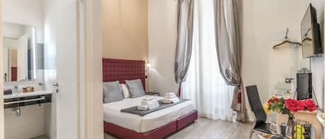 Habitación de hotel con cama, mesita de noche, espejo, zona de ducha y una pequeña mesa con un florero.