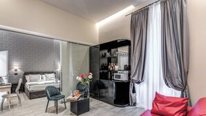 Habitación de hotel moderna con cama, escritorio, silla, pequeña mesa con un florero, sofá rojo, zona de cocina y un ventanal con cortinas.