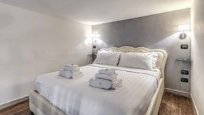 Una cama bien hecha con sábanas y toallas blancas, un cabecero, lámparas de noche y suelo de madera.