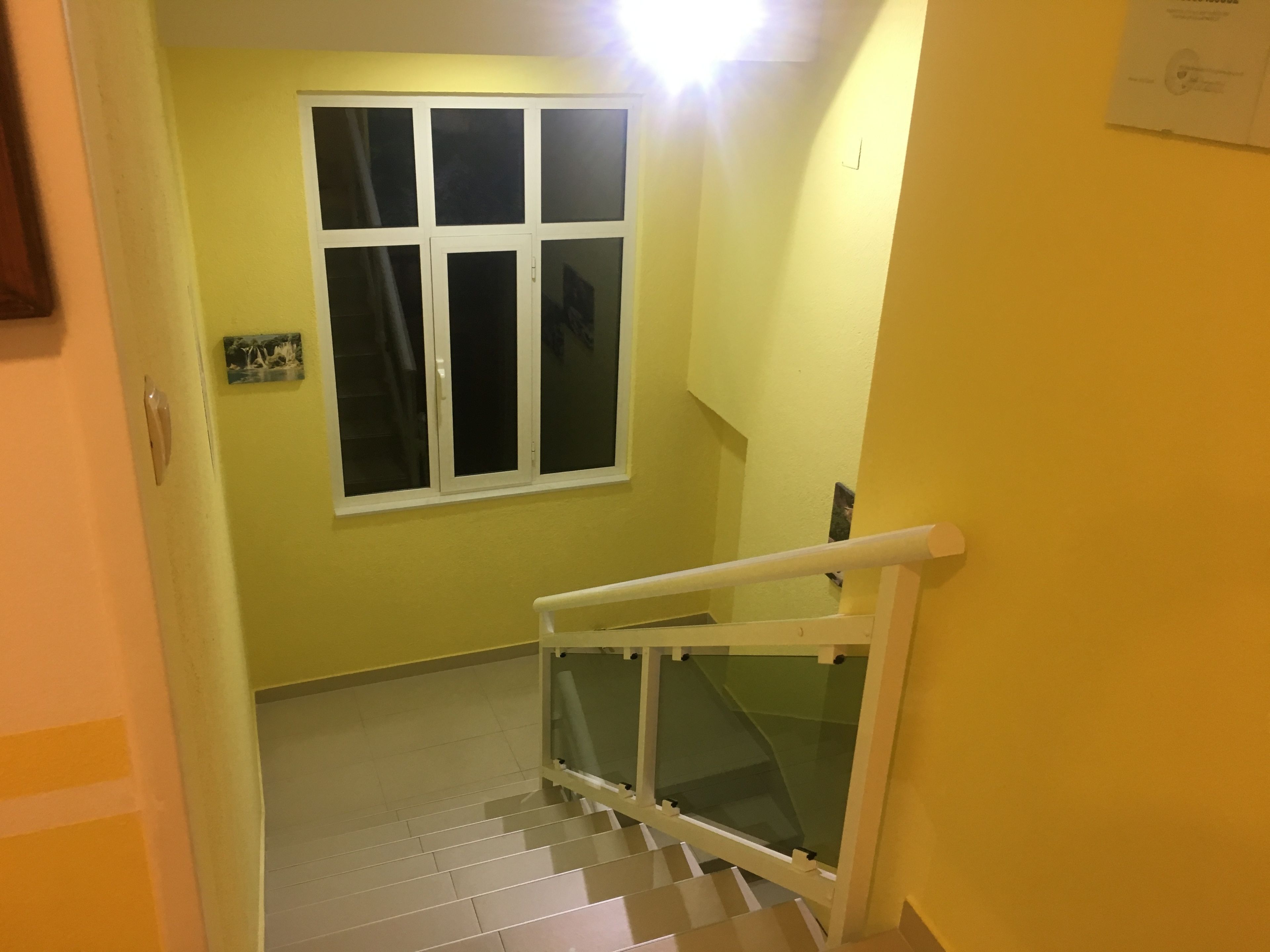 Una escalera con barandilla blanca y paneles de vidrio que conduce a una habitación con ventana y una lámpara.
