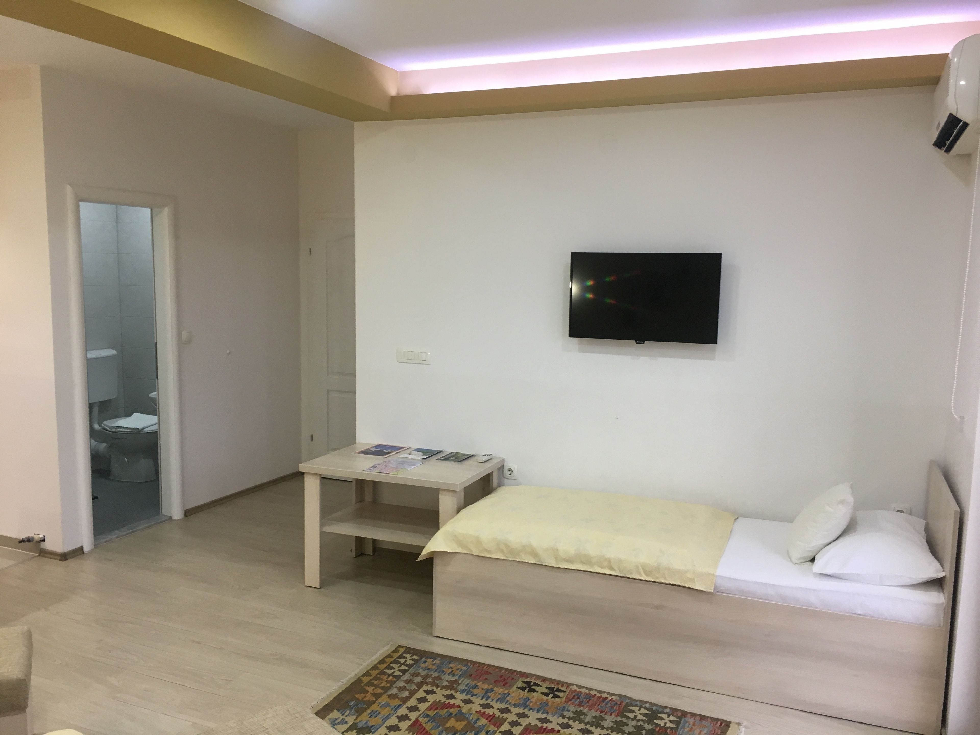A small, well-lit room with a bed, a TV mounted on the wall, and a small table with magazines.