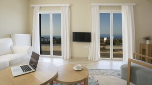 A modern living room with a white sofa, a laptop on a wooden table, a flat-screen TV mounted on the wall, and a view of a beach through large sliding glass doors. - Boo Premium Living Villas (Lefkada)