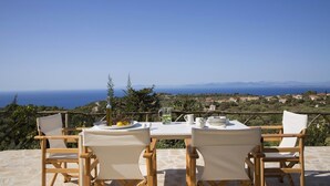 A table set with plates, a bottle, and lemons, overlooking a coastal landscape with houses and mountains. - Boo Premium Living Villas (Lefkada)