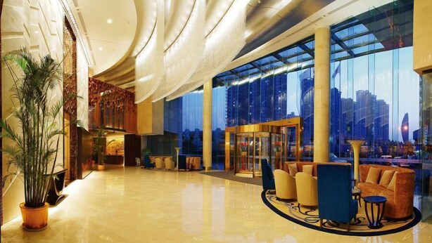 A spacious lobby with a high ceiling, large windows offering a city view, a reception desk, and seating areas. - Cenbest Jinling Grand Hotel (Wuhu)